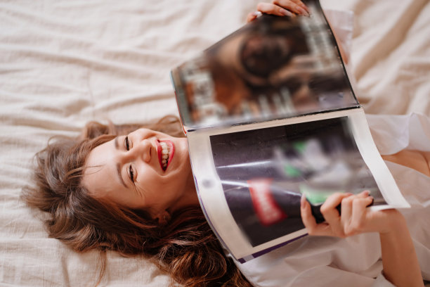 一个迷人而快乐的女人躺在床上看杂志。图片下载