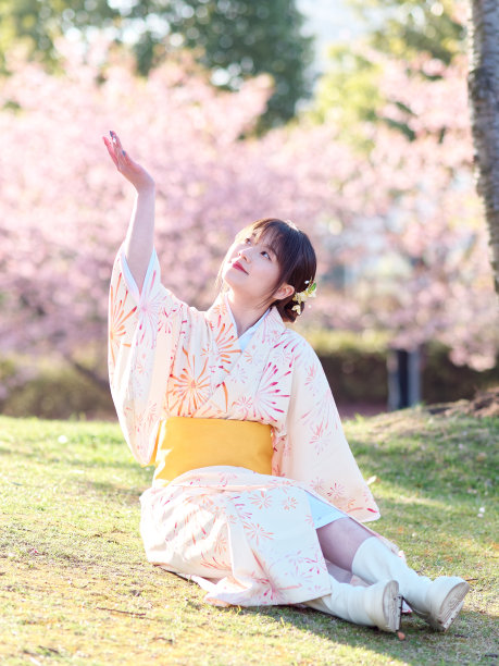 美丽的年轻中国女孩在日本和服坐在草地上，樱花盛开的背景在春天的花园，美丽，情感，生活方式，表达和人的概念。图片下载