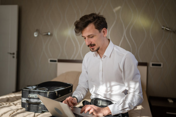 一名成年白人商人坐在酒店房间的床上，用笔记本电脑工作，准备会议或远程在线会议，真人复制空间，穿着白衬衫图片下载