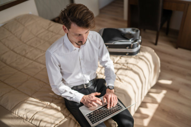 一名成年白人商人坐在酒店房间的床上，用笔记本电脑工作，准备会议或远程在线会议，真人复制空间，穿着白衬衫图片下载