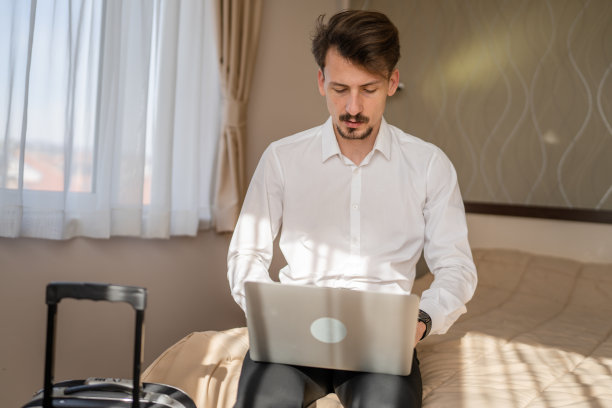 一名成年白人商人坐在酒店房间的床上，用笔记本电脑工作，准备会议或远程在线会议，真人复制空间，穿着白衬衫图片下载