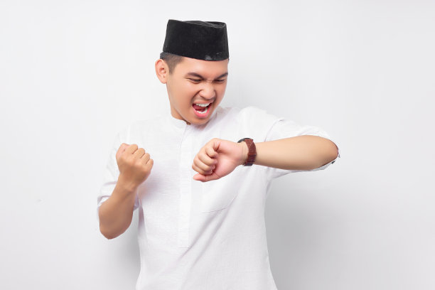 身穿阿拉伯服装的年轻亚裔穆斯林男子兴奋地举起手臂，看着手表，在白色背景上庆祝成功。人们信奉伊斯兰教的生活方式图片下载