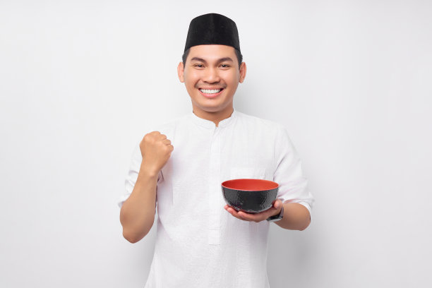 兴奋的年轻亚洲穆斯林男子穿着阿拉伯服装站着，拿着碗说，是时候开斋了，孤立在白色背景上。人们信奉伊斯兰教的生活方式图片下载