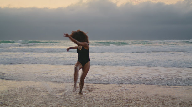 女孩表演者在多云灰色的天空跳舞海滩。女人在海洋中移动身体。图片下载