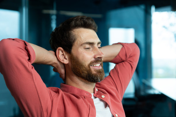 一个年轻的男性程序员，自由职业者，在办公室辛苦了一天后，正在办公桌前休息。他坐在椅子上，闭上眼睛，双手放在脑后，冥想，梦想，微笑图片下载