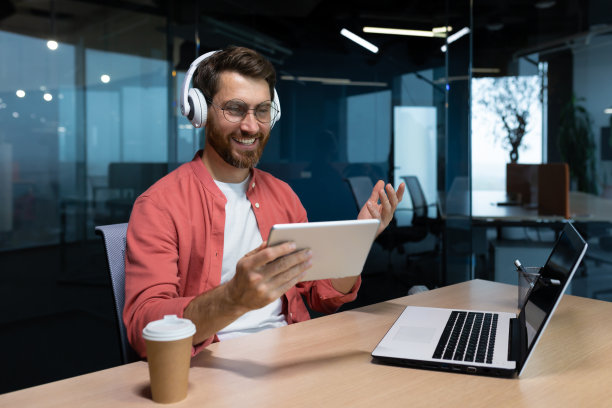 一个年轻的商人、自由职业者、学生、程序员坐在办公室的桌子旁，耳机里戴着一台笔记本电脑，手里拿着一台平板电脑。视频通话，听音乐图片下载