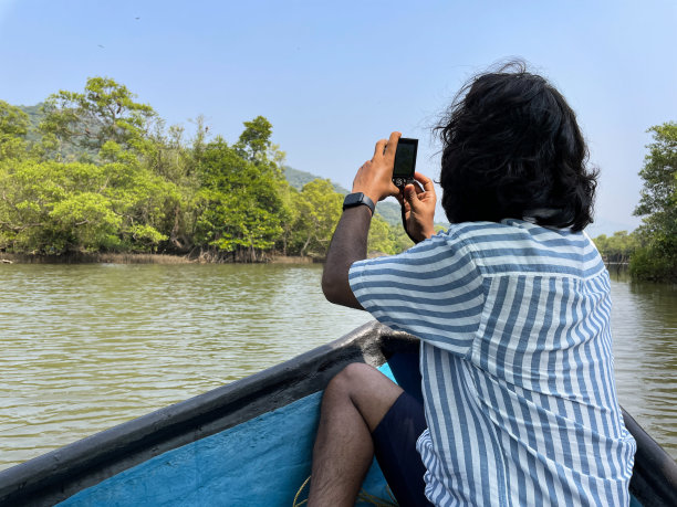 印度男子坐在渔船船尾用数码相机拍照的图像，船在泻湖浅水漂流，热带岛屿天堂，带棕榈树的林地，后视图，焦点在前景图片下载