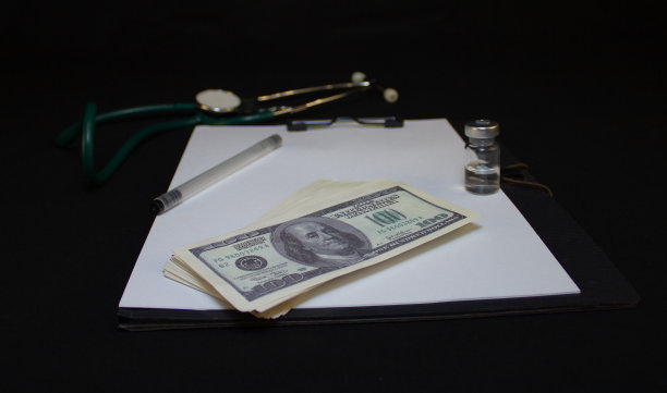 平板上放着一瓶药，旁边是钞票、美元和听诊器。黑色背景上摆放着用于接待病人和收集病历的医疗设备。图片下载