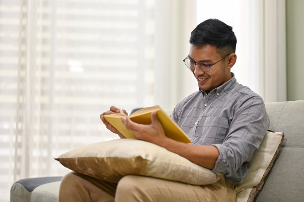 快乐的亚洲男人穿着便服，戴着眼镜，在客厅的沙发上看书。图片下载