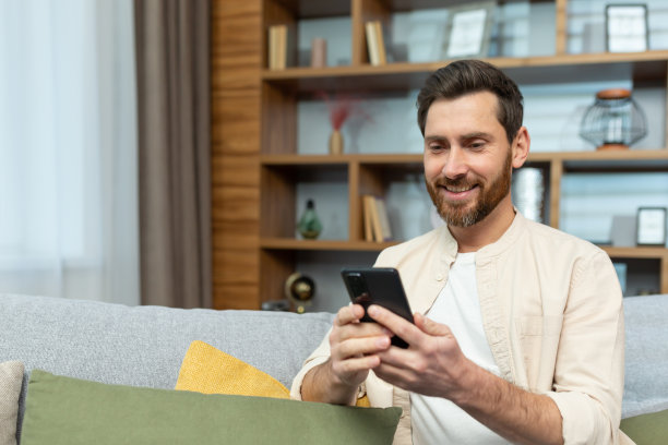 家里一个穿衬衫留着胡子的成熟男人正坐在客厅的沙发上，打着电话，发着短信，上网浏览图片下载