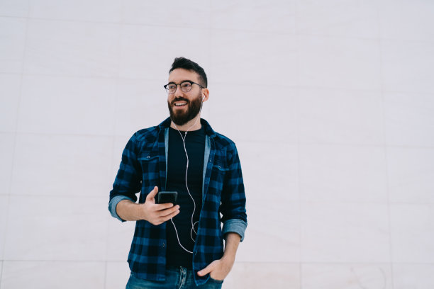 无忧无虑的潮人戴着光学眼镜使用手机媒体应用程序，在宣传区域微笑，快乐的男性戴着耳机享受积极的广播音乐，靠近宣传背景和复制空间图片下载