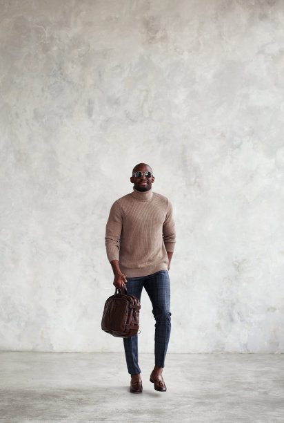 时尚的黑人男士走在灰色的墙上，穿着时髦的衣服，棕色的皮包和乐福鞋，高领毛衣。时尚风格图片下载