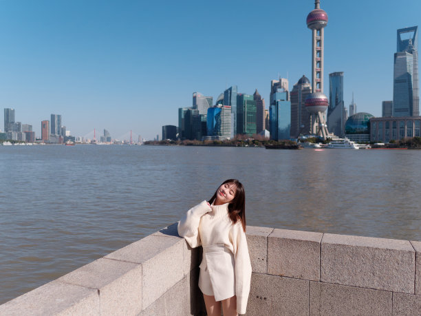 美丽的年轻女子，黑色长发，白色裙子，眼睛闭上，模糊上海外滩地标建筑背景在阳光灿烂的日子。情感、人、美、旅行、生活理念。图片下载