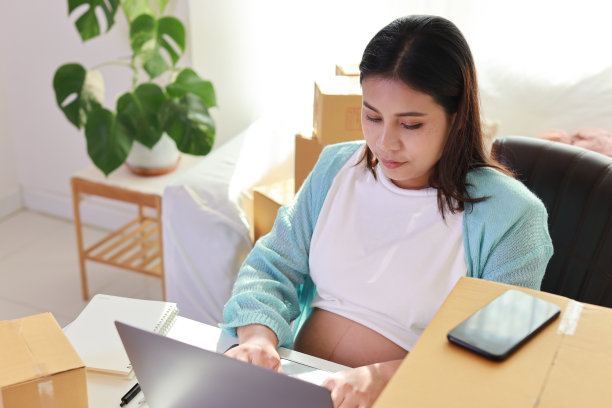 有吸引力的快乐亚洲孕妇坐着，使用移动智能手机，用电脑为客户订购。准妈妈在怀孕期间为孩子的出生做准备和工作图片下载