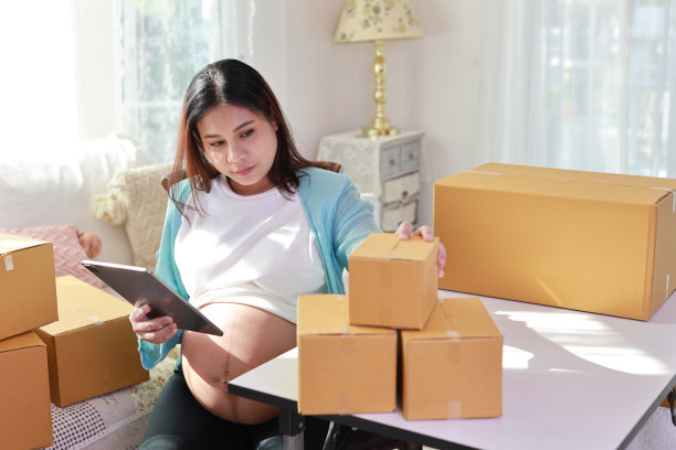 有吸引力的快乐亚洲孕妇坐着，使用移动智能手机，用平板电脑为客户写订单。准妈妈在怀孕期间为孩子的出生做准备和工作。图片下载