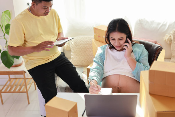 亚洲情侣一同进行盒子包装，年轻孕妇坐着使用手机写客户订单，丈夫在一旁工作图片下载