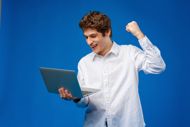 年轻人站在衬衫，拿着笔记本电脑，看着相机在蓝色的背景图片下载