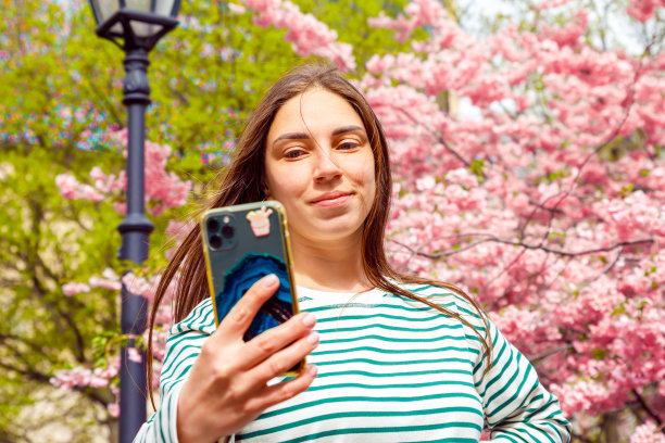 女子用手机视频通话，自拍。年轻微笑的欧洲白人女子在樱花樱花的背景，户外公园。图片下载