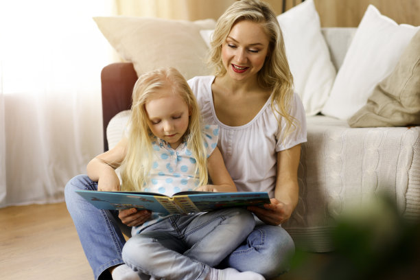 幸福的家庭。金发年轻的母亲读书给她可爱的女儿，坐在木地板在阳光明媚的房间。母亲的概念图片下载