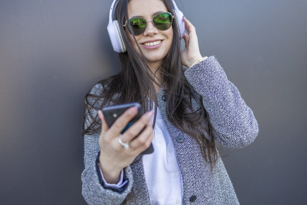 美丽的女人戴着耳机在她的耳朵上听音乐在她的手机。图片下载