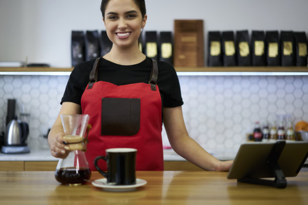 快乐的白人女性20多岁的咖啡师，穿着制服站在咖啡馆的酒吧里，用玻璃过滤器制作香气咖啡饮料，微笑着繁荣的女咖啡师，用罐子和杯子工作图片下载
