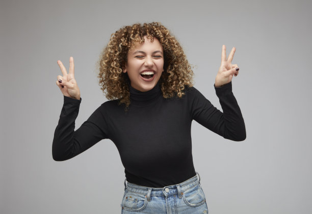 年轻女子与卷发微笑，同时闭上眼睛，使胜利的标志。图片下载