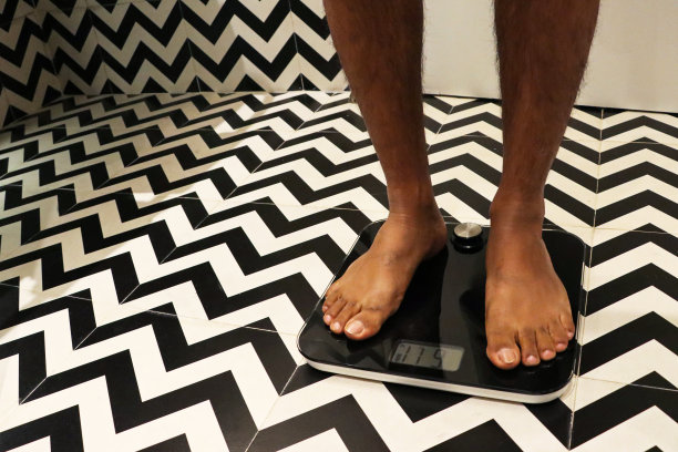 特写照片，裸足的无法辨认的人站在秤前的白色浴室门，豪华酒店浴室单色之字形图案的墙壁和地砖，关注前景，节食和饮食失调的概念图片下载