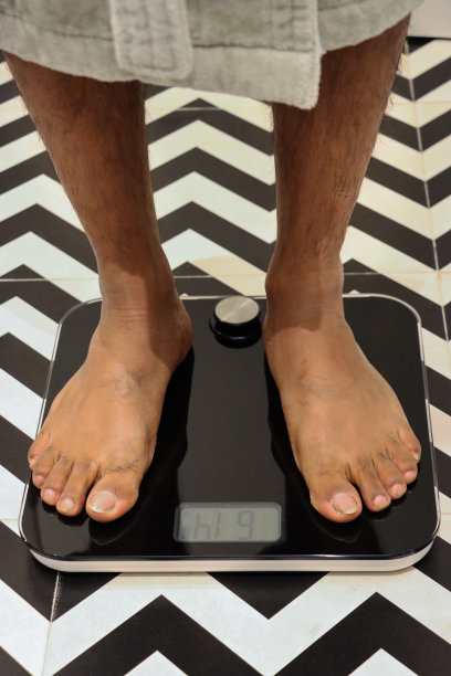 全画幅脚无法识别的人穿着浴袍站在秤，豪华酒店浴室单色之字形图案的墙壁和地砖，关注前景，节食和饮食失调的概念图片下载