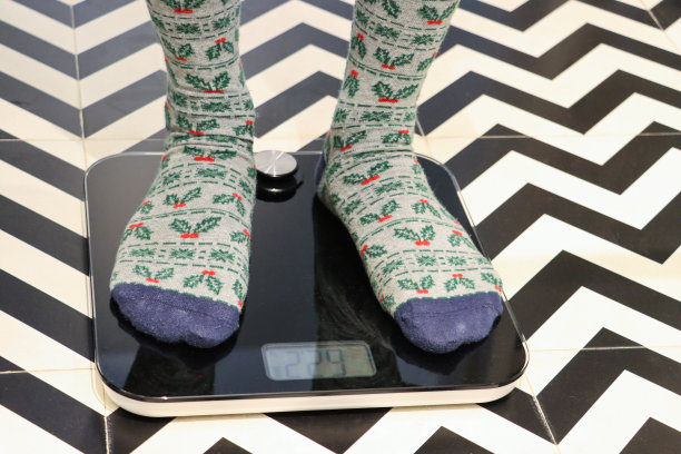 特写图像，无法识别的人穿着冬青叶和浆果设计的圣诞图案袜子站在秤上，豪华酒店浴室单色之字形图案地砖，专注于前景，节食和饮食失调的概念图片下载