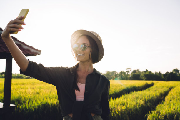 在普吉岛参观稻田时，快乐的潮博主对着手机摄像头微笑，开朗的女视频博主用智能手机应用程序拍摄搞笑视频内容图片下载