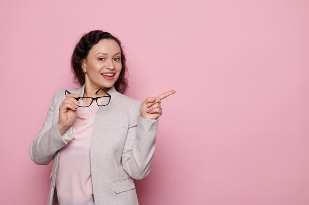 可爱的女人在灰色优雅的西装，拿着时尚的眼镜，看着相机，指着复制空间在粉红色的背景图片下载