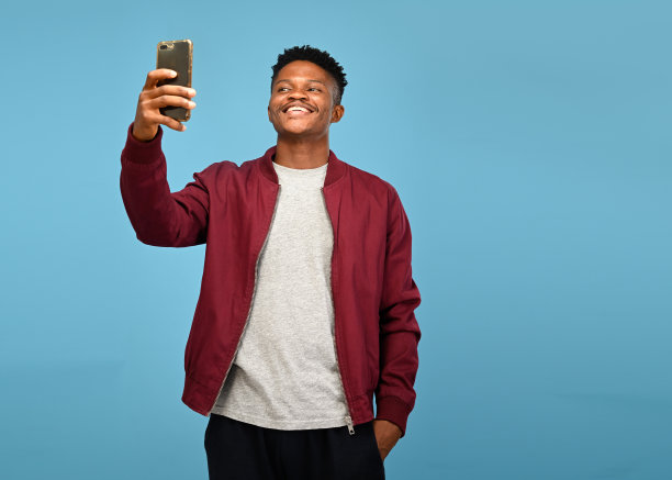 年轻英俊的男子站在他的手拿着他的智能手机在他的头上，把他放在口袋里，时尚地看着他的智能手机图片下载