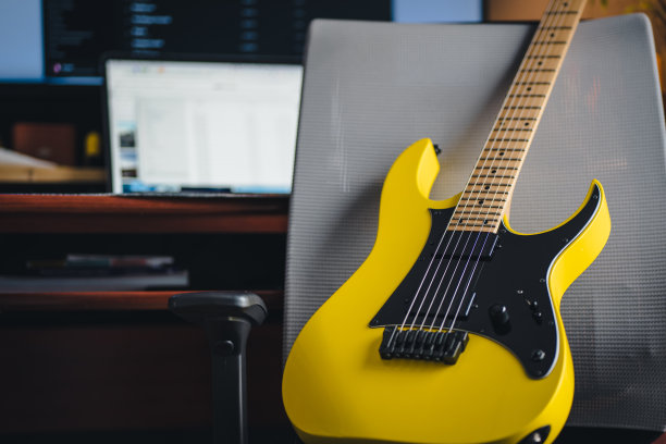 黄色电吉他站在办公室灰色椅子上，模糊的办公桌和笔记本电脑背景在一张库存照片中图片下载