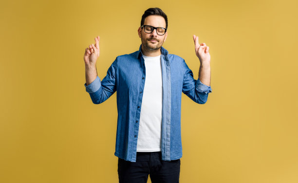 迷人平静的年轻成年男子穿着蓝色牛仔衬衫闭上眼睛，交叉手指好运孤立在黄色背景图片下载