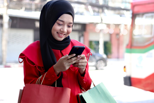 快乐的微笑年轻的亚洲穆斯林妇女戴着头巾，穿着红色连衣裙，用手机打字短信，拿着购物袋走在步行街，美丽的购物走在城市道路上的肖像图片下载