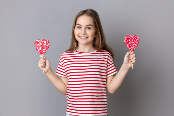 小女孩可口的心形棒棒糖，表情俏皮，微笑着看着镜头。图片下载