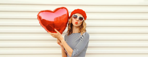快乐的年轻女子的肖像与红色心形气球吹她的嘴唇送甜蜜的飞吻戴贝雷帽在白色背景图片下载