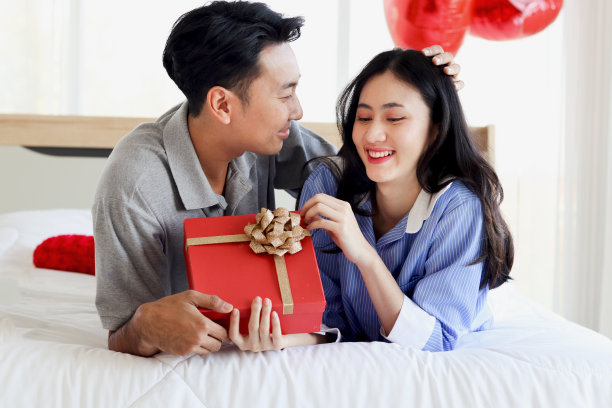 幸福的亚洲情侣躺在卧室的白色床上看着对方，浪漫的男人给他的女朋友送红色的情人节礼物，庆祝难忘的情人节纪念日图片下载