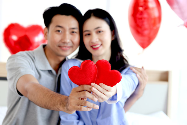 幸福的亚洲情侣在卧室抱着红心，向镜头展示，浪漫的恋人在白色的卧室庆祝难忘的情人节纪念日，爱的庆祝概念。图片下载