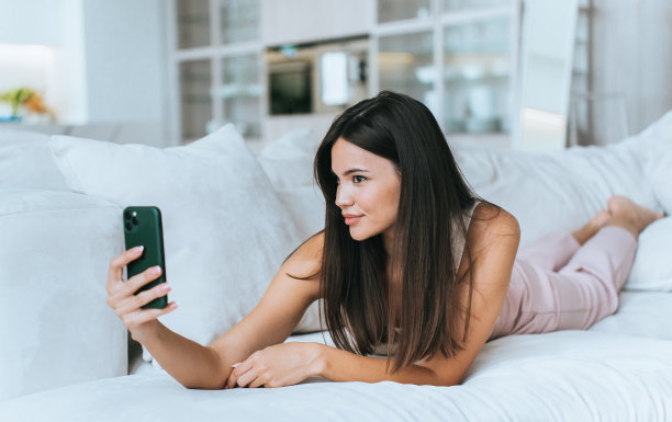 可爱的亚洲女孩在浅粉色裤子躺在沙发上拿着电话视频电话回家。黑发女博主通过互联网与订阅者谈话。时尚模特女子在酒店放松。图片下载