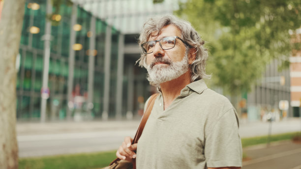 一个头发灰白、胡子花白的中年男人走来走去，环顾四周。成熟的绅士戴着眼镜，肩上背着包，在城市景观背景下走过广场图片下载