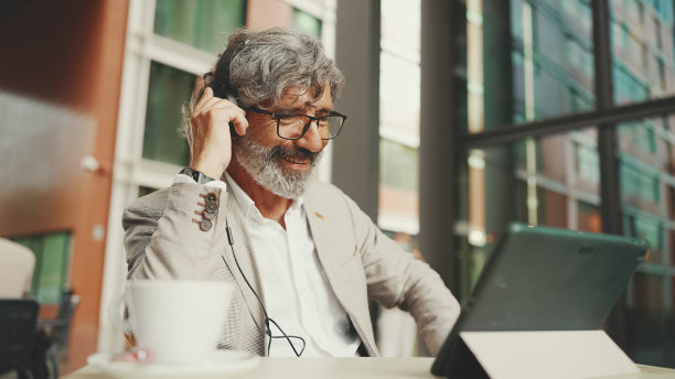 成熟的商人，留着胡子，戴着眼镜和耳机，坐在户外咖啡馆里用平板电脑工作图片下载