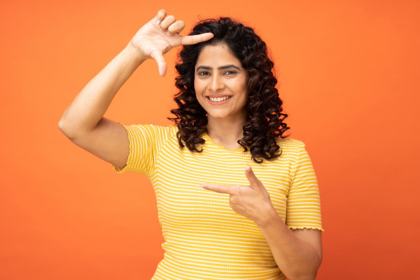 有吸引力的漂亮的现代漂亮卷发快乐的年轻女子显示与框架手势隔离在明亮的橙色背景，股票照片图片下载
