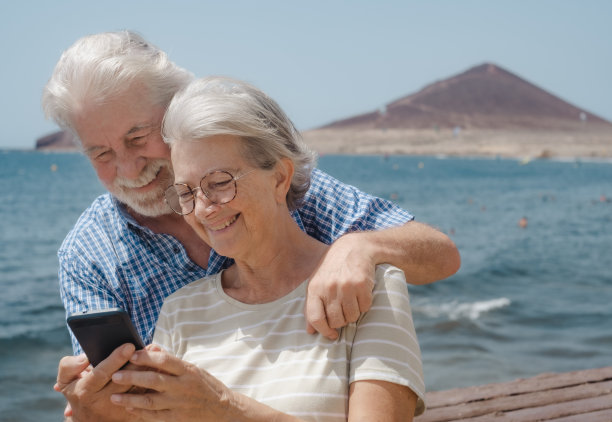 在阳光明媚、风大的天气里，享受海上度假的老年人在海边用手机拥抱的美丽的白种人夫妇图片下载