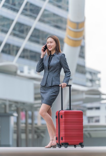 年轻优雅的亚洲女商人，穿着全套办公套装，带着红色的行李和购物袋去出差。边走边用手机聊天。图片下载