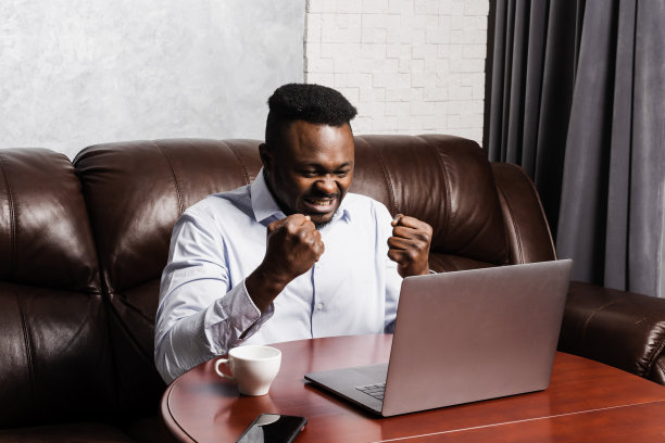 快乐的非洲男子坐在笔记本电脑前为获胜而高兴。快乐的非裔美国人与笔记本电脑举起手，微笑着看着收入的收入数额和喜悦。图片下载