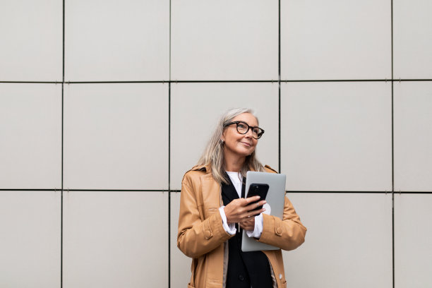 时尚的灰色头发成熟的女商人与手机和笔记本电脑的背景墙的建筑图片下载