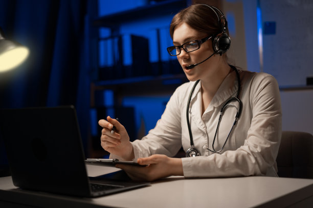 一个快乐微笑的年轻医生在办公室在笔记本电脑前在晚上头戴耳机的肖像。图片下载