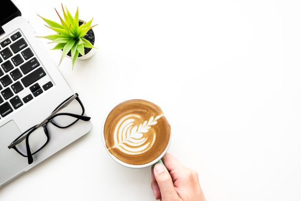 手持一杯拿铁咖啡的俯视图在白色的办公桌桌上，笔记本电脑和办公用品。顶视图与复制空间，平铺。图片下载