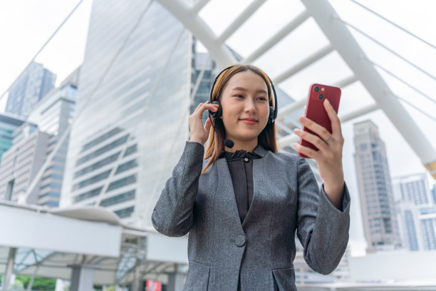 在商业区，一位年轻的亚洲女商人穿着灰色的办公套装，用智能手机和她的客户通过耳麦交谈，脸上带着自信的微笑图片下载